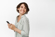 © Cookie Studio - Portrait of young smiling woman holding smartphone, turn head at copy space, chatting cellular, standing against white background