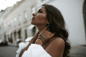 Engaging young lady with brunette wavy hair, tanned body in white stylish blouse, puffed sleeves, posing outdoors clasping neck 