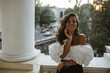 Pretty young lady with long brunette hair, tanned body and closed eyes in white off-shoulder top, metallic bracelets standing on balcony against street view background in evening