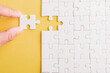 © sorapop - Top view flat lay of hand-holding last piece white paper jigsaw puzzle game last pieces put to place for solve problem complete mission, studio shot on a yellow background, quiz calculation concept