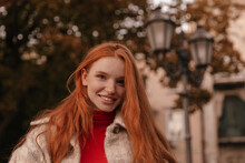 Close-up Portrait Of Charming Young Girl Smiling And Posing Outdoors. Pretty Lady With Smooth Long Red Hairstyle, Freckles And Trendy Clothes, Looking Straight Forward In City