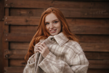 Wall Mural - Beauty portrait of young foxy lady against wood background. Charming long-haired girl with freckles, nude makeup, plaid coat, smiling, holding collar and posing outdoors