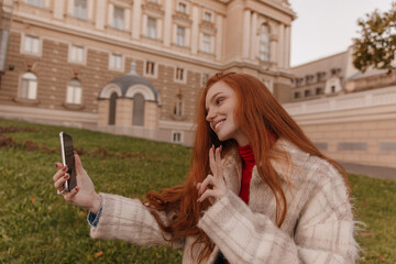 Wall Mural - Pretty young ginger making selfie in autumn city. Adorable red-haired girl wearing plaid coat, showing peace with fingers and smiling