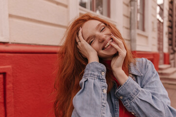 Wall Mural - Joyful young foxy lady smiling and posing outdoors. Cute ginger with smooth long hair, freckles, denim shirt, touching head and narrowing eyes against bright background