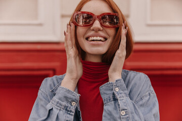 Wall Mural - Close-up portrait of joyful young ginger posing against bright colourful background outdoors. Adorable girl with red turtleneck, denim shirt and trendy sunglasses, smiling and touching head