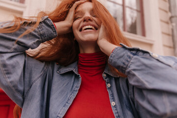 Wall Mural - Lifestyle portrait of young ginger moving on street. Cute long-haired girl with freckles, red turtleneck and denim shirt, smiling, touching head and posing outdoors