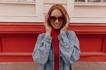 Wall Mural - Lifestyle portrait of young trendy girl adjusting sunglasses on street. Stylish red-haired lady with denim shirt and modern accessories, smiling and posing outdoors against bright background