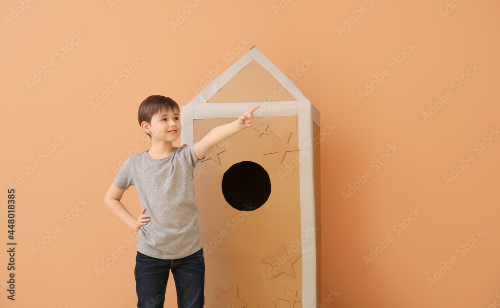 Cute little boy with cardboard rocket pointing at something on color background