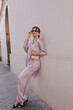 © Look! - Fashion portrait of young lady leaning on light wall. Attractive short-haired brunette wearing silk pastel outfit, trendy sunglasses with chain, black shoes, holding little bag and posing outdoors
