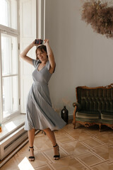 Lifestyle portrait of young lady at beautiful furnished studio. Adorable girl with short hair, trendy blue dress and black shoes, posing full-length, holding phone and smiling against vintage sofa