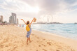 Happy female tourist walks barefoot on a sandy beach in the JBR area of Dubai and admires the panoramic view of the Ain Ferris Wheel