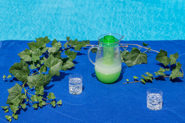 A blue poolside beach towel is used as a tablecloth topped with two glasses with ice cubes and sparkling water and a pitcher of grapefruit juice with ivy leaves