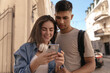 © Look! - Close-up portrait of pretty young pair in sunny town. Cheerful dark-haired lady with white headphones and blue clothes looking at phone near attractive friend wearing beige t-shirt