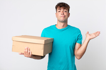 Wall Mural - young handsome man feeling puzzled and confused and doubting and holding a cardboard box