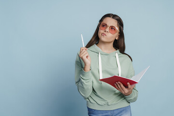 Wall Mural - Cute teenage girl in sunglasses holding a notebook, writing something down. on a blue background, copy space