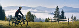 Man riding bicycle on grassy hill and looking at beautiful misty mountains. Male bicyclist enjoying panoramic view of majestic mountains during bicycle ride. Concept of sport, bicycling and nature.