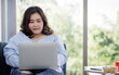 © Bangkok Click Studio - Asian young happy beautiful plus size sexy fat chubby smart female model sit relaxing on sofa smiling look at camera using laptop computer work from home in quarantine time from covid pandemic moment