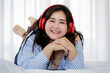 © Bangkok Click Studio - Asian young happy calm fat overweight woman lay down with happy smiling deep emotion peacefully while listening to music from big red earphones use playlist from black tablet at home in bedroom