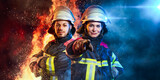 Fototapeta Sport - Ein Feuerwehrmann und eine Feuerwehrfrau stehen zusammen vor Feuer und Blaulicht