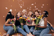 Group of diverse women with bouquets having fun under falling petals. Six happy females sitting together against a brown background.