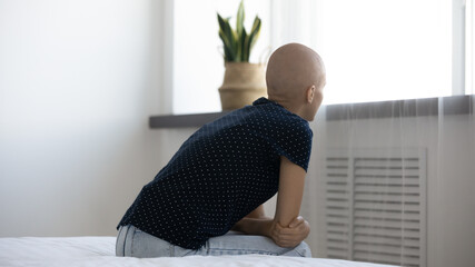 Wall Mural - Rear back view stressed young hairless after chemotherapy woman sitting on bed looking in distance, thinking of oncology treatment or operation, feeling depressed alone indoors, cancer fight concept.