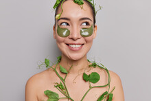 Hygiene And Personal Care Concept. Healthy Young Asian Woman With Clean Skin Has Toothy Smile Applies Collagen Patches Wrapped In Green Plant Removes Dark Circles Isolated Over Grey Background