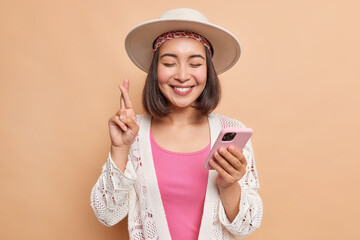 Wall Mural - Horizontal shot of cheerful dark haired young Asian woman with happy smile keeps fingers crossed for good luck holds mobile phone isolated over beige background anticipates good news results.