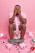 © WHstudio Leushin N - Dark skinned man poses comfortably on toilet bowl feels shy hides face with hands surrounded by party accessories isolated over pink background. Male model in wc. Hygiene defecation concept.