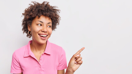 Wall Mural - Photo of smiling curly haired Afro American woman points index finger right shows discount sale banner wears casual pink t shirt isolated over white background demonstrates cool awesome product