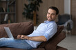 Young man using laptop. Portrait of smiling successful guy recline on pillow on comfy couch with pc surf information at web chat online. Happy man remote student hold notebook on laps look at camera