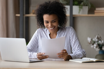 Wall Mural - Happy young African American woman sit at desk at home office work online on computer with paperwork documents. Smiling biracial female read good news in paper correspondence. Success concept.