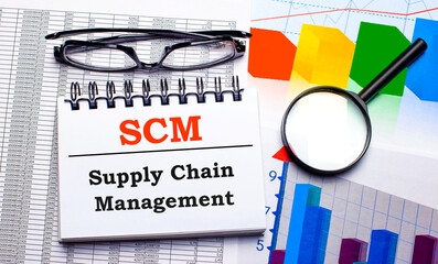 On the desktop are glasses, a magnifying glass, color charts and a white notebook with the text SCM Supply Chain Management. Business concept. View from above