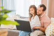 © Maria Moroz - Happy family, daughter sitting on her father's lap on couch at home looking at laptop monitor together.