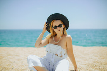 Wall Mural - Fashion photo of a girl in black glasses with a chain and a large straw hat, against the backdrop of the sea or ocean. Woman sunbathes on the beach, summer photo. Blonde with long hair