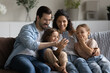 © fizkes - Happy family couple and little kids using mobile phone, sitting together on couch at home, taking selfie, making video call. Parents and children looking at smartphone screen, laughing