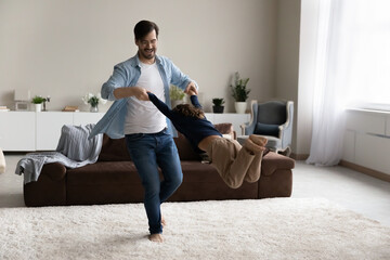 Wall Mural - Happy father and excited little son enjoying funny active energetic games in living room, playing swirl and sway. Dad spinning around, holding arms of kid flying in air, having fun at home.