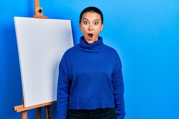 Poster - Beautiful hispanic woman with short hair standing by painter easel stand scared and amazed with open mouth for surprise, disbelief face