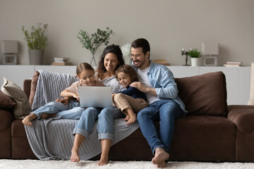 Happy millennial parents and gen Z kids relaxing on couch at home, using app on laptop, watching movie online, making video call, shopping on internet, playing virtual game on digital device