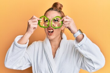 Sticker - Young blonde woman wearing bathrobe holding green pepper over eyes angry and mad screaming frustrated and furious, shouting with anger looking up.