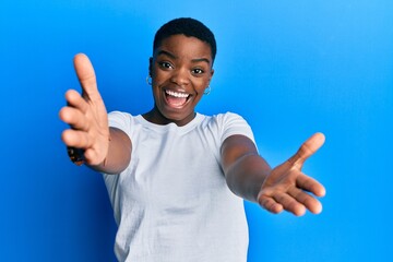 Wall Mural - Young african american woman wearing casual white t shirt looking at the camera smiling with open arms for hug. cheerful expression embracing happiness.