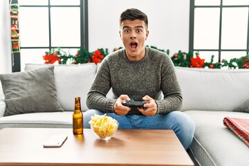 Sticker - Young hispanic man playing video game on christmas scared and amazed with open mouth for surprise, disbelief face