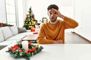 Wall Mural - Arab young man sitting on the table by christmas tree doing ok gesture shocked with surprised face, eye looking through fingers. unbelieving expression.