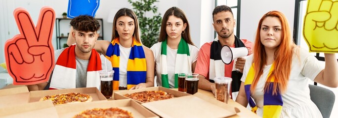 Wall Mural - Group of young hispanic people eating pizza supporting soccer team at home thinking attitude and sober expression looking self confident