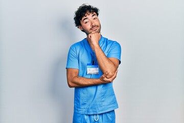 Poster - Young hispanic man wearing blue male nurse uniform looking confident at the camera with smile with crossed arms and hand raised on chin. thinking positive.