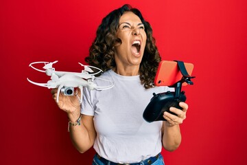 Sticker - Middle age hispanic woman using drone angry and mad screaming frustrated and furious, shouting with anger looking up.