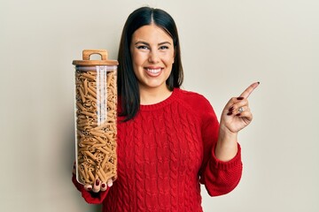 Poster - Beautiful brunette woman holding jar with macaroni pasta smiling happy pointing with hand and finger to the side