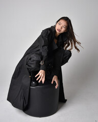 Full length portrait of pretty young asian girl wearing black tank top, utilitarian  pants, leather boots and large dark coat. Sitting pose with gestural hand movements,  isolated on  studio backgroun