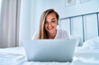 © Goffkein - Young happy smiling joyful millennial girl lying on a bed and using laptop with wireless headphones for watching video online, learning language courses, social networking and video calling at home