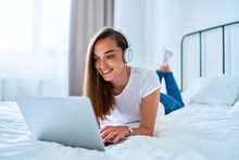 Young Happy Smiling Joyful Millennial Girl Lying On A Bed And Using Laptop With Wireless Headphones For Watching Video Online, Learning Language Courses, Social Networking And Video Calling At Home