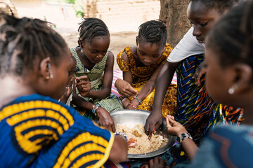 group of coulorfully dressed black girls sitting around a big metal bowl, sharing a typical african 